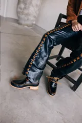 Pantalón de cuero de tiro medio y silueta recta levemente acampanada, color azul marino, con cierre frontal y recortes laterales intervenidos con pasacintas entrelazadas en contraste.