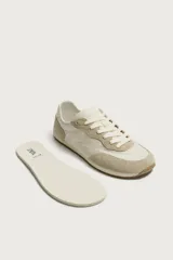 Zapatilla deportiva estilo barefoot, color marrón, con diseño de paneles en combinación de materiales y detalles en serraje. Cuenta con puntera redondeada y ancha, cordones en color crema y suela fina de goma flexible en contraste.
