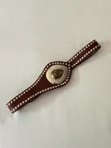 Cinto faja de cuero marrón con tachas metálicas plateadas en los bordes. Presenta una pieza central metálica con textura martillada y un dije en forma de gota con piedra natural.