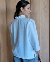 Camisa de manga tres cuartos color beige con cuello con volados y botones frontales. Presenta un detalle de color negro en la parte superior de las mangas.