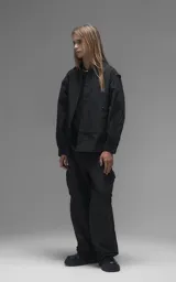 Chaqueta tipo overshirt de microfibra, color negro, con calce oversize y ruedo curvo. Presenta cierre frontal con broches a presión, bolsillos frontales de gran tamaño y cierres laterales a lo largo de las mangas para ajustar su amplitud.