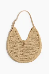 Cartera de hombro tipo hobo confeccionada en paja de papel trenzada de color beige, con asa regulable de cuero sintético y cierre de bucle con botón de nudo decorativo.