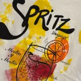 Remera blanca de corte clásico con estampa frontal ilustrada que representa un cóctel Spritz, incluyendo texto alusivo y detalles en tonos amarillos, naranjas y rojos con efecto de pinceladas.