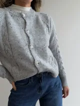 Cárdigan de punto gris con efecto jaspeado, diseño de trenzas en las mangas, cuello redondo y cierre frontal con botones. Presenta puños y ruedo acanalados.