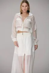 Chaqueta corta de organza en color blanco, con diseño semitransparente, cierre frontal con cremallera, cuello clásico y dos bolsillos frontales con solapa.