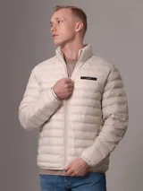 Campera acolchada color beige claro, con cuello alto y cierre frontal con cremallera. Tiene bolsillos laterales y logo de Calvin Klein en el pecho.