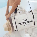 Bolso tipo tote bag de lona color beige con detalles de ribetes negros. Presenta el texto 'THE TOTE BAG' y 'LINCOLN'S' estampado en el frente. Cuenta con doble asa superior y correa ajustable extraíble.