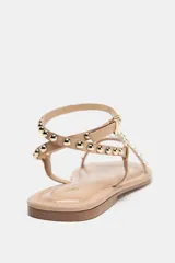 Sandalias planas color beige con tiras finas y pulsera al tobillo, decoradas con tachas metálicas doradas.