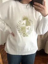 Buzo de punto liviano color blanco, con cuello redondo, mangas largas y diseño holgado. Presenta un bordado frontal en tono dorado con el escudo nacional de Uruguay.