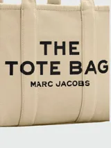 Bolso tipo tote de gran tamaño en color beige, confeccionado en lona resistente. Presenta el logo estampado en negro en el frente y detalles de cintas laterales con inscripciones. Cuenta con doble asa superior para llevar al hombro.