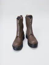 Botas de cuero color marrón de caña alta, con plataforma gruesa de goma y cierre metálico frontal con tirador. Presentan costuras a contratono y detalle de etiqueta de la marca en la lengüeta.