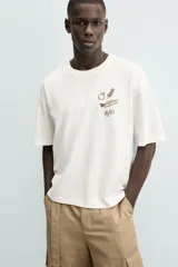 Camiseta de manga corta color blanco roto, corte relajado, con un pequeño bordado/estampado en el pecho que incluye una manzana, una rama de laurel, una trompeta y dos dados.