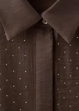Camisa de satén color marrón chocolate, con cuello camisero, cierre frontal de botones ocultos y mangas largas con puños abotonados. Presenta un diseño recto adornado con pequeños detalles de strass brillantes distribuidos por toda la prenda.