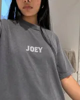 Remera oversize gris oscuro con corte holgado, mangas cortas y estampa trasera en blanco y negro con forma de estampilla que muestra a una persona con un gato y el texto 'Kaboa Industries'.