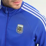 Campera deportiva Adidas de la selección argentina, color azul con cierre frontal, cuello alto, puños y cintura acanalados. Presenta las icónicas tres tiras blancas en las mangas, el logo del trébol de Adidas en el pecho derecho y el escudo de la AFA en el pecho izquierdo.