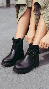 Bota corta de cuero negro con suela track de goma, detalle de tira cruzada con hebilla metálica lateral y paneles de gamuza en contraste.