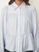 Camisa de corte amplio y estilo romántico, color celeste, con cuello clásico, cierre frontal de botones y diseño de canesú fruncido. Presenta mangas largas abullonadas con puños elásticos.