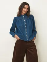 Camisa de jean de corte recto con cuello alto tipo mao con volados, cierre frontal con botones a presión y mangas largas con hombros ligeramente abullonados.