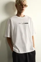 Camiseta de corte holgado con cuello redondo y manga corta. Presenta un diseño frontal con la frase New York Essence combinando tipografía cursiva y letras en bloque con efecto relieve.
