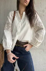 Camisa de inspiración boho de color crudo, con mangas largas abullonadas y puños decorados con detalles bordados geométricos en tonos tostados y dorados. Presenta cuello en V con cierre frontal de botones.