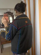 Chaqueta oversize de paño de lana en color beige, con costuras a la vista bordadas a mano en crochet con hilo multicolor en tonos rojos, azules, amarillos y verdes. Presenta cuello camisero y diseño amplio.