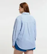 Camisa plus size alargada confeccionada en tela anarruga a rayas verticales celestes y blancas, con cuello inglés, botones delanteros para cierre y mangas largas con puños abotonados.