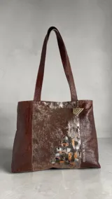 Cartera de cuero natural en color marrón jaspeado, tipo tote bag, con diseño amplio y asas dobles para el hombro. Presenta intervención artística pintada a mano en el frente con detalles metalizados y geométricos, y dije metálico colgante.