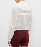 Campera cortavientos deportiva modelo cropped, color blanco traslúcido, con cierre frontal de cremallera, capucha estructurada, puños elásticos y bajo ajustable con cordón.