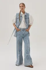 Chaleco largo de jean celeste con cuello camisero, sin mangas, con bolsillos frontales, laterales e internos, y lazo ajustable en la cintura.