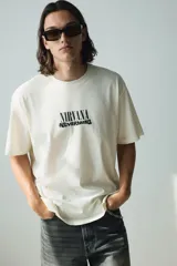 Remera color crema de corte holgado con estampado frontal del logo de la banda Nirvana y el título de su álbum Nevermind en color negro.
