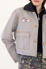 Campera corta de jean color visón, con cuello desmontable de tela engomada, cierre frontal y bolsillos plaqué con solapa en el pecho. Cuenta con un parche bordado en el delantero.