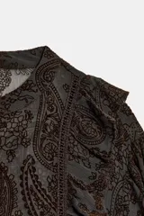 Blusa de manga larga con cuello redondo, confeccionada en tejido semitransparente con estampado paisley en tonos marrones oscuros. Presenta detalles de volantes y apliques de terciopelo en el frente y hombros, con cierre frontal de botones forrados.