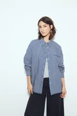 Camisa de silueta amplia y caída fluida, color celeste con rayas verticales blancas. Presenta cuello camisero clásico, cierre frontal con botones y un bolsillo aplicado en el pecho. Las costuras están resaltadas en azul oscuro.