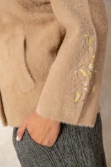 Campera de textura afelpada en color beige, con corte recto, cuello camisero y cierre frontal oculto. Presenta un botón joya ornamental en el cuello y un bordado artesanal en una de las mangas.