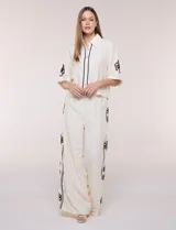 Conjunto de dos piezas en color marfil, compuesto por una camisa de manga corta con cuello camisero y cierre frontal de botones con ribetes negros, y un pantalón palazzo de corte amplio con líneas laterales negras. Ambas prendas cuentan con detalles bordados en contraste.