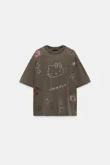 Remera oversize de color marrón grisáceo con efecto lavado, cuello redondo y manga corta. Cuenta con múltiples aplicaciones de brillos con motivos de Hello Kitty, estrellas, cerezas y moños.