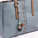 Cartera tipo tote de cuerina color celeste, con detalles y asas de mano en color tostado. Presenta un llavero decorativo colgante con cuentas de colores, costuras decorativas frontales y una correa ajustable y desmontable.