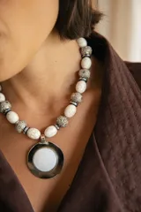 Collar corto con cuentas de piedra reconstituida blanca con vetas, cuentas tibetanas decoradas y cuentas de hematite. Presenta un medallón central circular con centro de nácar y borde plateado. Incluye cadena de acero quirúrgico con ganchito para regular el largo.
