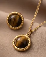 Set compuesto por un collar con cadena fina y dije circular, y un par de aros tipo botón a juego. Ambas piezas presentan una piedra natural ojo de tigre engarzada en una estructura de metal dorado con borde trenzado.