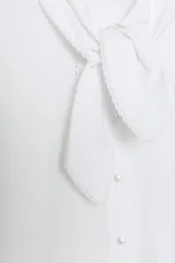 Camisa de popelín blanca con cuello solapa decorado con puntilla y lazo frontal. Presenta mangas largas con puños abotonados y cierre frontal con botones.