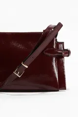 Bandolera de charol color bordó, con cierre superior y correa ajustable. Presenta detalles de trabillas decorativas en los laterales con herrajes metálicos dorados.
