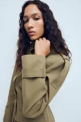 Chaqueta de estilo militar en color beige, con cuello subido, cierre frontal de botones, bolsillos de plastrón con solapa en el frente y detalle de hombreras con trabillas.