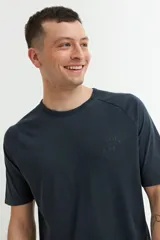 Remera de manga corta color azul oscuro o negro deslavado, con cuello redondo y corte raglán. Presenta un pequeño estampado en relieve o tono sobre tono en el pecho con texto y un símbolo.