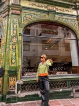 Camisa de manga larga en color verde oliva, con cuello babero en tono contrastante y detalle de volado en el ruedo en color naranja. Presenta cierre frontal con botones y mangas abullonadas con puños ajustados.