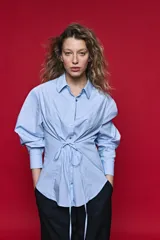 Camisa blanca de manga larga con volumen y estructura, cierre frontal con botones y lazo ajustable en la cintura que realza la silueta.