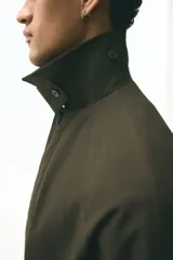 Gabardina estilo Mac de color marrón oscuro, confeccionada en sarga suave. Presenta cuello clásico, cierre frontal con botones ocultos, manga raglán con trabilla y botón en los puños, bolsillos insertados y cinturón extraíble para atar a la cintura. Tiene una abertura en la espalda y forro interior.