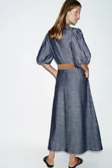 Vestido midi de denim azul con cuello camisero, mangas abullonadas y cierre frontal oculto con botones. Incluye un cinturón marrón con hebilla metálica.