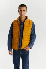 Chaleco puffer acolchado color mostaza, con cuello alto y cierre frontal. Diseño ligero y versátil con bolsillos laterales.