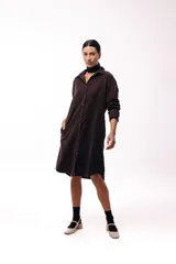 Vestido camisero de color marrón oscuro, con cuello clásico, cierre frontal de botones y mangas largas. Presenta un diseño de corte recto y largo midi.