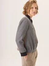 Chaqueta tipo bomber con diseño estructurado, confeccionada en una combinación de lana gris jaspeada y paneles laterales en color beige. Presenta cuello, puños y ruedo en tejido elástico negro, cierre frontal con cremallera y bordado sutil en el pecho.